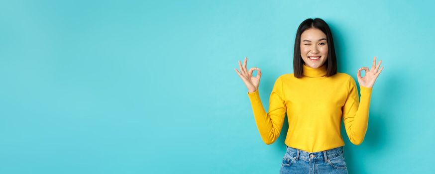 Cheerful asian female model showing okay gestures, smiling and looking impressed, praise product, standing over blue background.