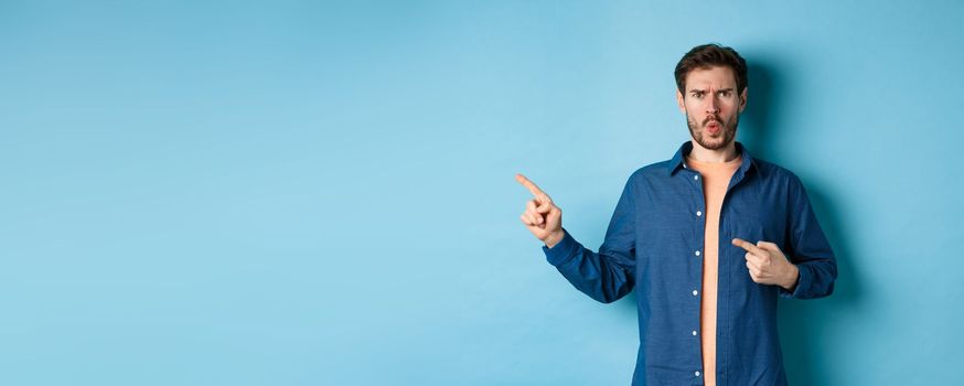 Shocked and offended young man frowning and looking hurt, pointing fingers left at empty space, complaining on something, standing on blue background.