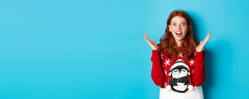 Winter holidays and Christmas Eve concept. Surprised redhead girl receiving good news, raising hands and gasping amazed, standing in awe over blue background.