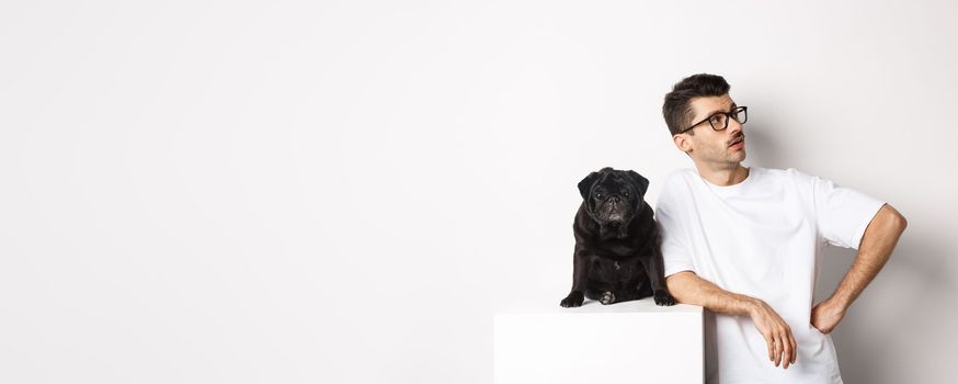 Handsome young man standing near cute black pug, looking right with arrogant expression, standing over white background.