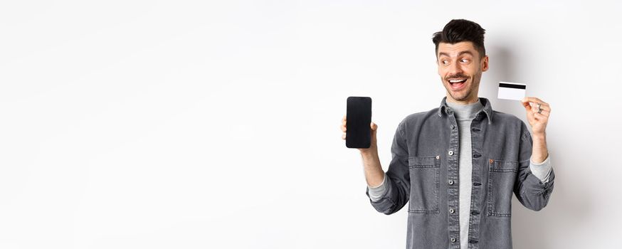 Handsome smiling man in denim jacket looking at empty smartphone screen and showing plastic credit card, good internet offer, standing against white background.