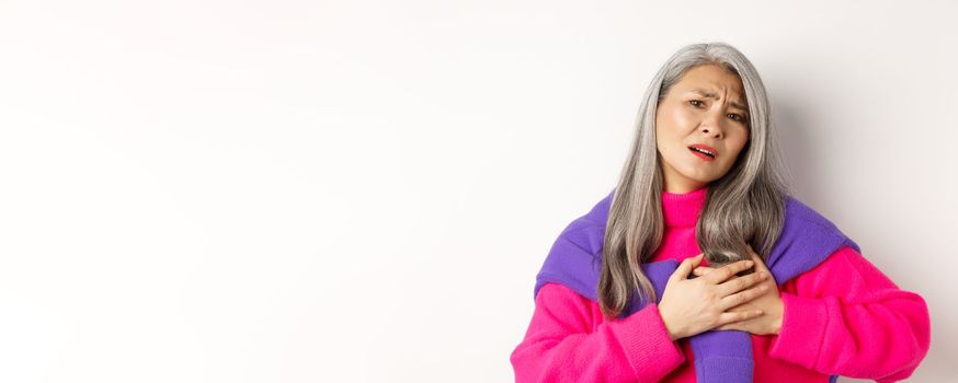 Worried asian mother holding hands on heart and grimacing, looking anxious and having pain in chest, feeling hurt, standing over white background.