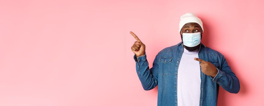 Coronavirus, lifestyle and global pandemic concept. Amused african-american male model in face mask pointing, looking left at promo offer, showing banner, pink background.