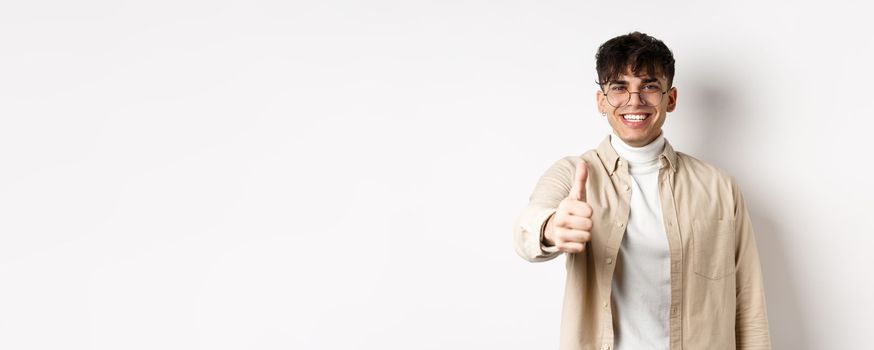 Image of happy young man showing thumb up and smiling satisfied, praise good job, say well done and look pleased, standing on white background.