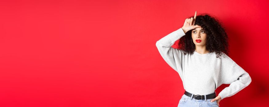 Arrogant woman with curly hair, showing loser sign on forehead and looking aside, mocking someone, standing on red background.