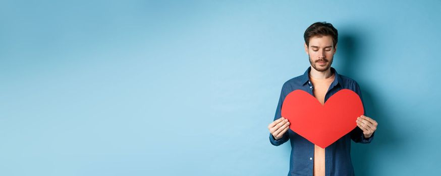 Lonely guy looking sad at valentines red heart with sad face, standing over blue background.