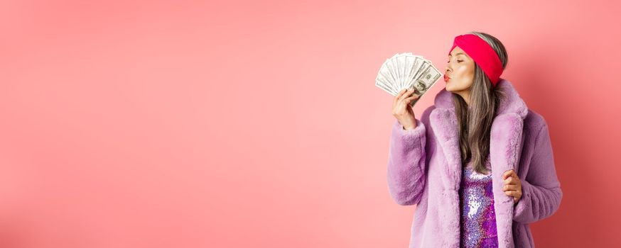 Shopping and fashion concept. Fashionable and rich senior woman kissing dollars money, looking pleased, wearing purple faux fur coat with party dress, pink background.