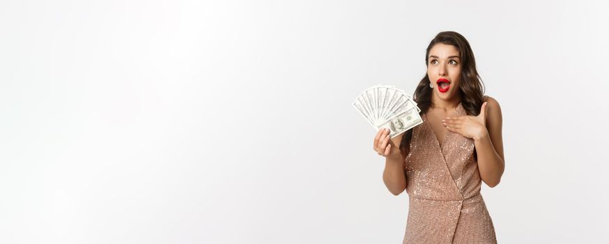 Shopping concept. Image of surprised and excited woman holding money, staring at upper left corner amazed, standing with dollars in glamour dress, white background.