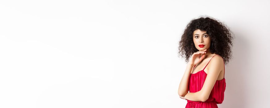 Beauty and fashion. Elegant lady with curly hair and red lips, fashionable makeup, wearing dress, gently touching chin and looking sensual at camera, white background.