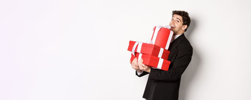 Concept of christmas holidays, celebration and lifestyle. Handsome man in black suit, holding pile of gifts and smiling, wishing happy new year, standing over white background.