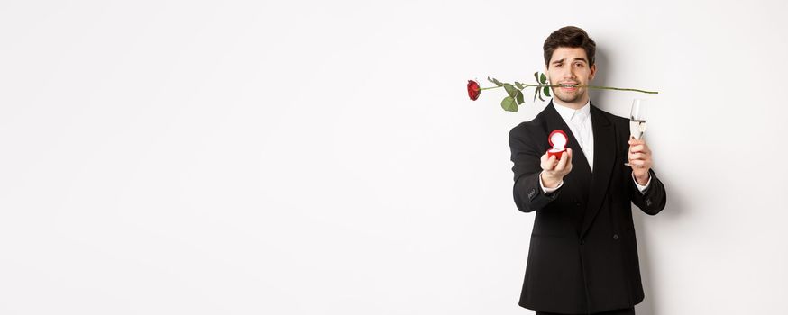 Romantic young man in suit making a proposal, holding rose in teeth and glass of champagne, showing engagement ring, asking to marry him, standing against white background.
