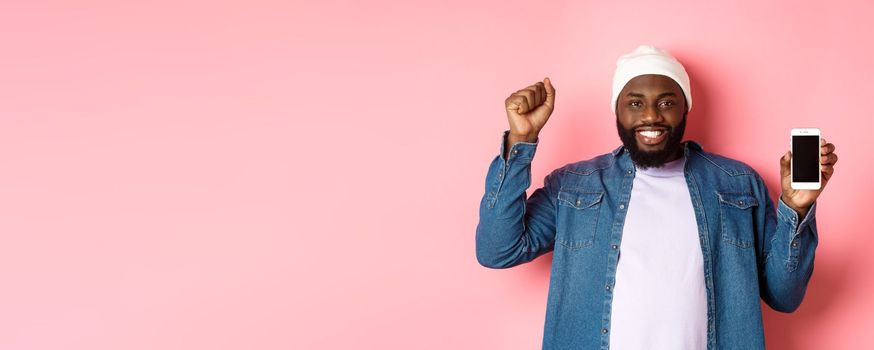 Online shopping and technology concept. Cheerful Black man rejoicing and showing mobile screen, raising hand up satisfied, triumphing while standing over pink background.