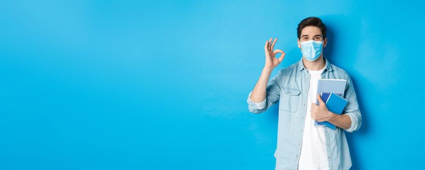 Education, covid-19 and social distancing. Guy student in medical mask looking happy, holding notebooks, showing ok sign, standing over blue background.