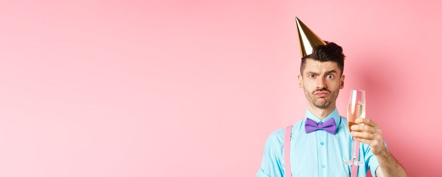 Holidays and celebration concept. Troubled young man in party hat, frowning with doubtful face, raising glass of champagne perplexed, standing on pink background.