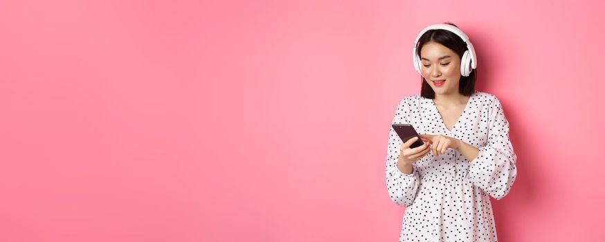 Beautiful asian woman texting message on smartphone, listening music in headphones, standing over pink background.