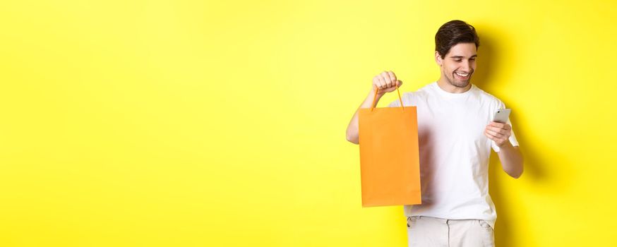 Concept of discounts, online banking and cashback. Happy guy showing shopping bag and looking satisfied at mobile screen, yellow background.