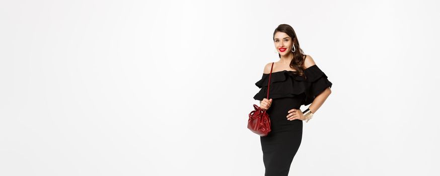 Beauty and fashion concept. Full length of elegant young woman going shopping in black dress, heels and purse, looking confident, standing over white background.