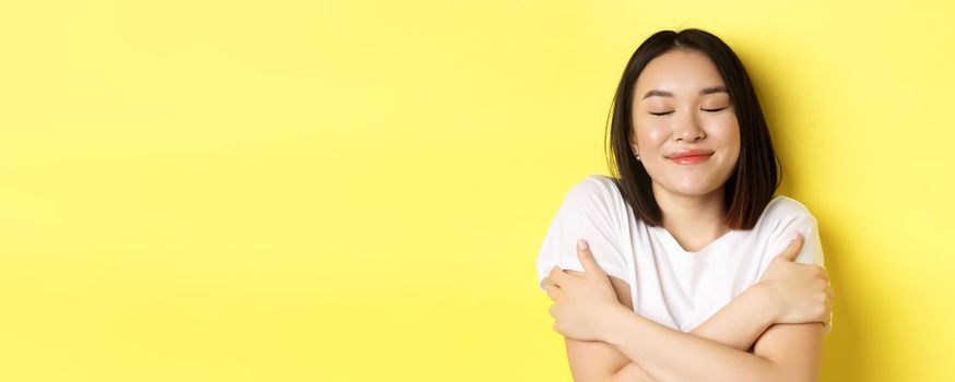 Close up of romantic asian girl hugging herself and dreaming, close eyes and smile while imaging something tender, standing over yellow background.