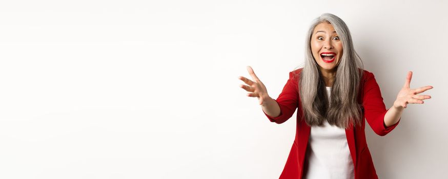 Surprised and friendly asian woman reaching hands forward and smiling, welcoming you, greeting someone, standing over white background.