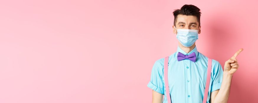 Covid-19, pandemic and health concept. Smiling caucasian man in medical mask pointing finger right, showing advertisement, standing on pink background.