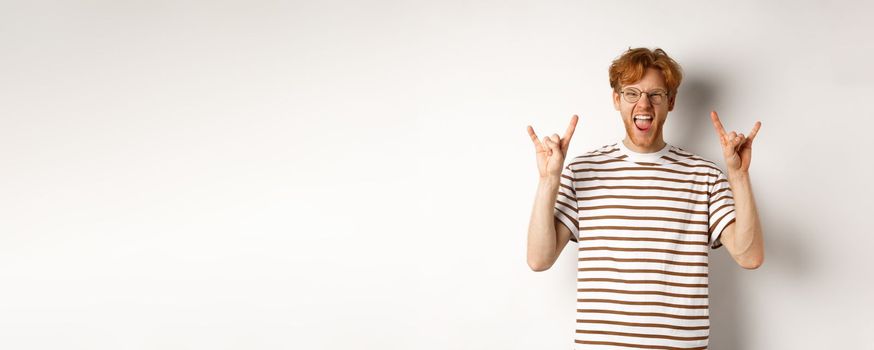 Funny and happy redhead man having fun, showing rock-n-roll horn and sticking tongue, enjoying party, standing over white background.