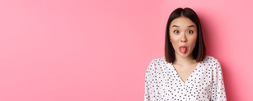 Beauty and lifestyle concept. Close-up of funny and cute asian woman showing tongue, staring at camera silly, standing over pink background.