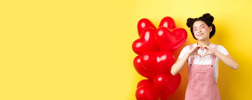 Valentines day concept. Cute asian girl dreaming of romance, close eyes and showing heart gesture, smiling happy, standing near red romantic balloons, yellow background.