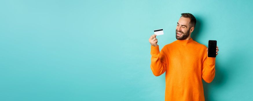 Online shopping. Satisfied man using credit card and showing mobile screen, looking pleased, standing over light blue background.