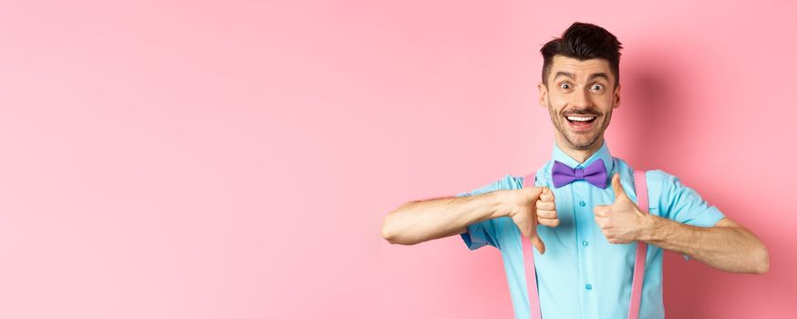 Funny young man smiling and showing thumbs up down, judging something and being indecisive, making choice, standing on pink background.