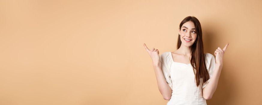 Dreamy caucasian girl making choice, pointing fingers sideways and looking at upper left corner pensive, picking product, standing on beige background.