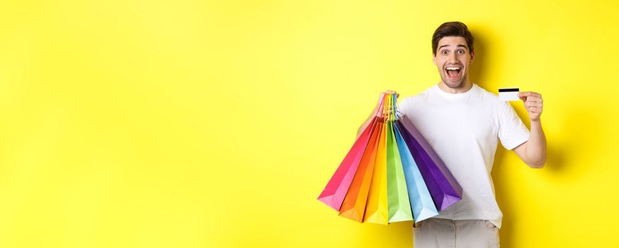 Happy attractive man holding shopping bags, showing credit card, standing over yellow background.