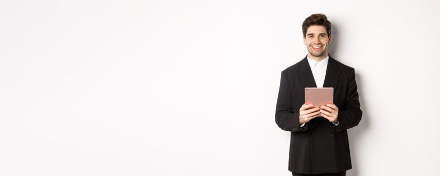 Image of handsome businessman in trendy suit, holding digital tablet and smiling, standing against white background.