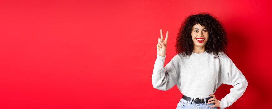 Young pretty lady in sweatshirt showing number two, making an order and smiling, standing on red background.