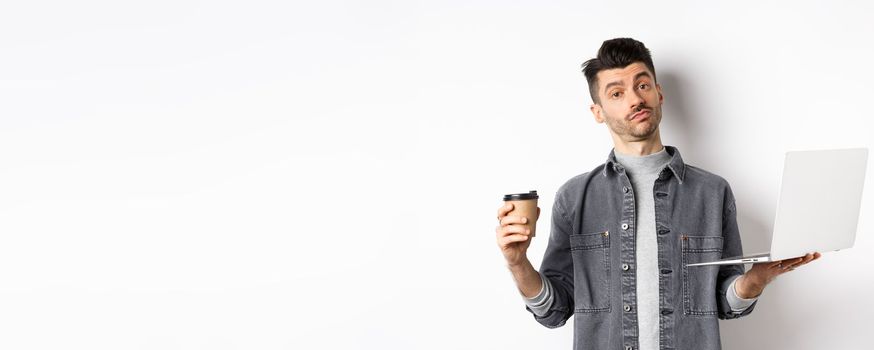 Handsome young man trying new coffee from cafe, holding cup and working on laptop, standing thoughtful on white background.