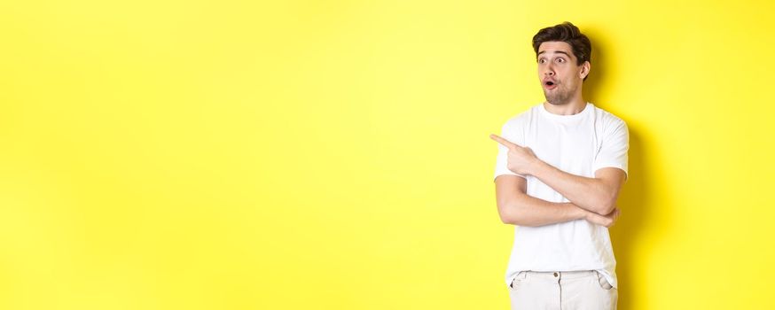 Impressed man in white t-shirt, looking and pointing finger left at promo, check out advertisement, standing against yellow background.