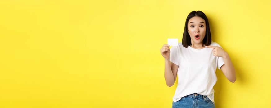 Young asian woman in casual white t-shirt showing plastic credit card and smiling at camera, yellow background.