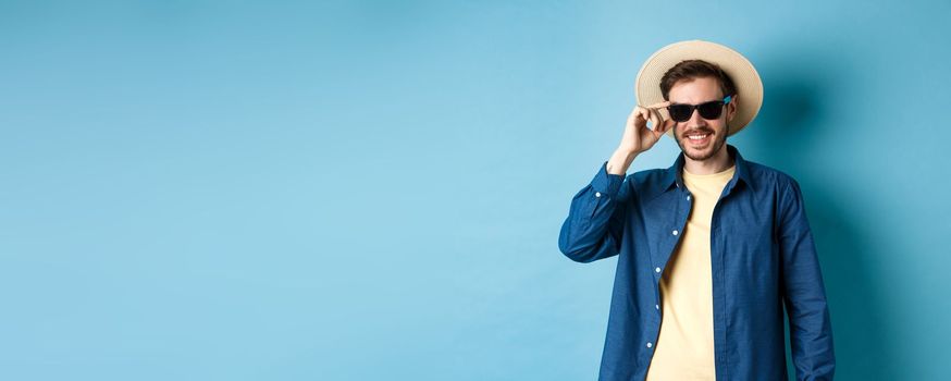 Happy smiling guy going on summer vacation, wearing straw hat and black sunglasses, standing on blue background.