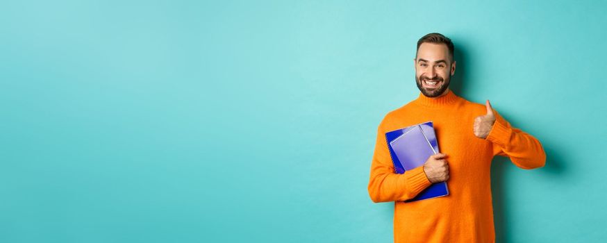 Education. Smiling bearded man holding notebooks and smiling, going on courses, showing thumbs-up in approval, like something, standing over turquoise background.