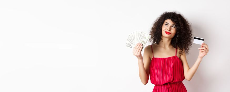 Shopping. Woman thinking and smiling, holding money with plastic credit card, wearing red elegant dress and evening makeup, white background.