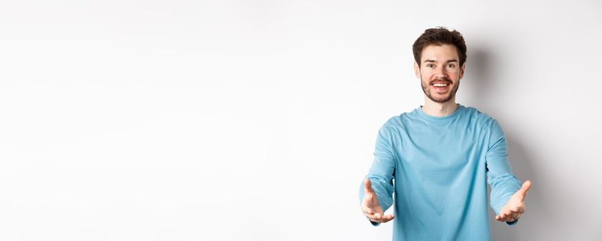Attractive caucasian guy in casual clothes stretch out hands to hold something, receiving and smiling, standing over white background.