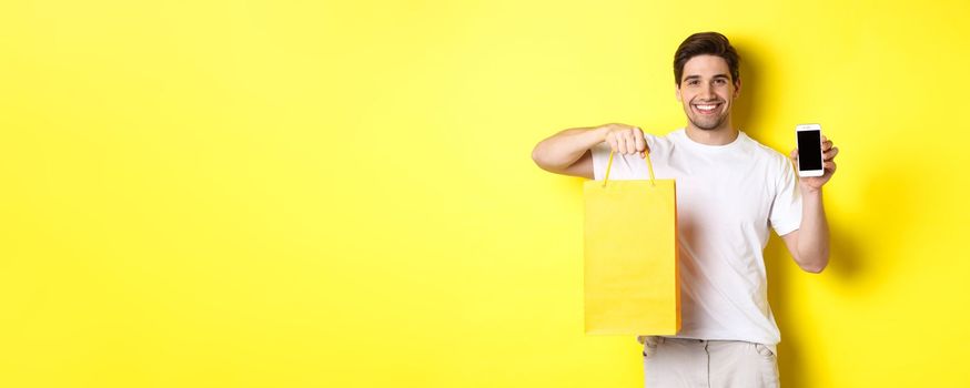 Concept of mobile banking and cashback. Young happy guy holding shopping bag and showing smartphone screen, yellow background.