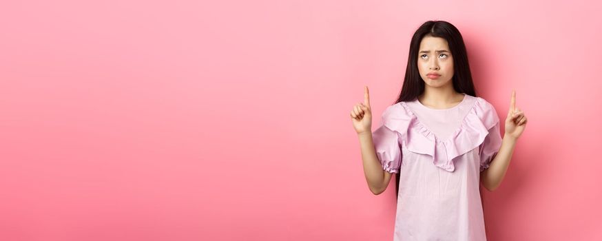 Sad pitiful asian girl sulking, pointing and looking up with disappointed face, feeling upset, standing against pink background.