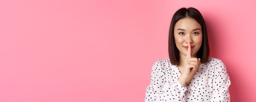 Close-up of mysterious asian woman hiding a secret, hushing and telling to keep quiet, standing over pink background.