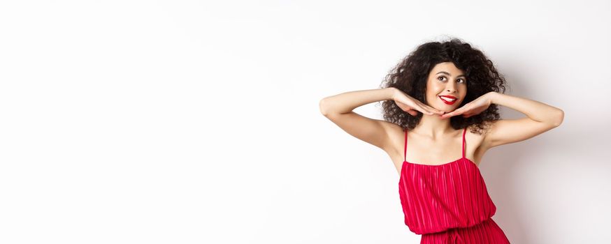 Beautiful lady with curly hair, wearing red dress, showing hear face with makeup and smiling, feeling carefree on white background.