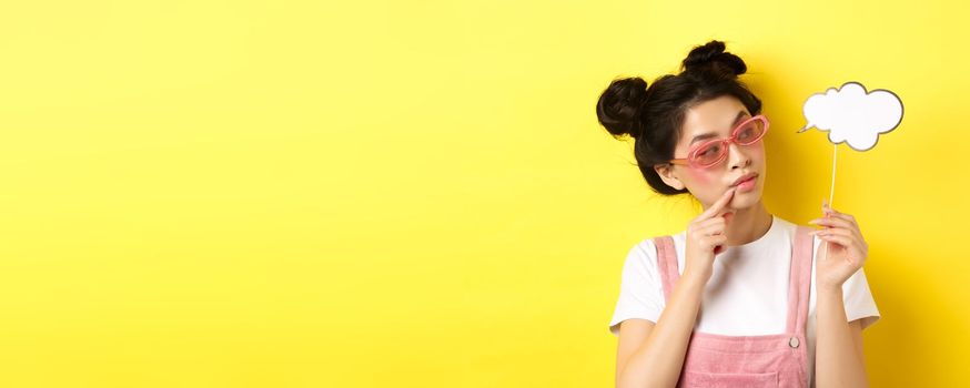 Summer and fashion concept. Stylish glamour asian girl in sunglasses, holding comment cloud party mask and looking aside, yellow background.