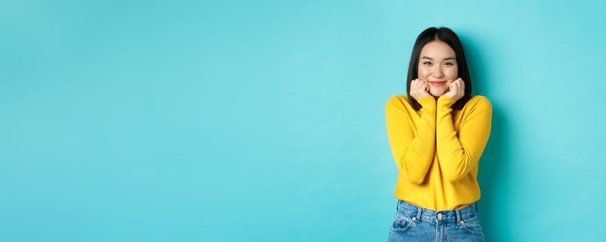 Beauty and fashion concept. Beautiful asian woman blushing and smiling, looking at something cute and silly, lean face on palms, standing over blue background.