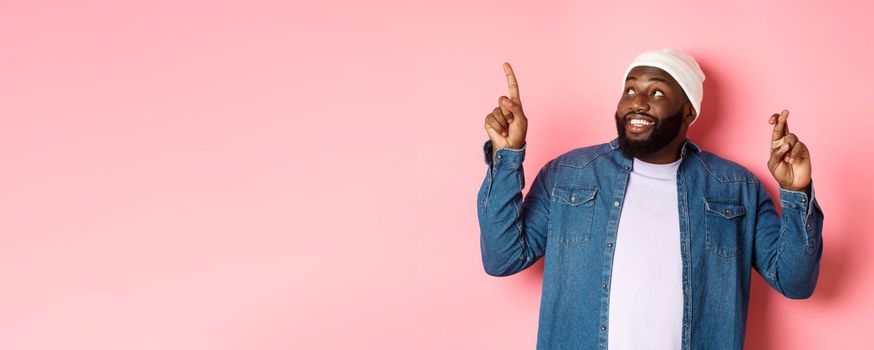 Hopeful african-american man making a wish, cross fingers for good luck, pointing and looking at upper left corner, dreaming about buying something, pink background.