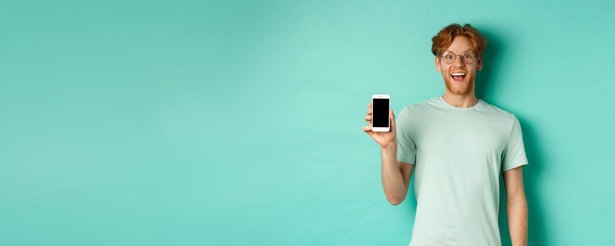 Online shopping. Cheerful redhead man in glasses and t-shirt showing smartphone screen and smiling, standing over mint background.