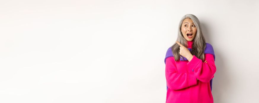 Portrait of amazed asian senior woman gasping fascinated, pointing at upper left corner, demonstrate special promotion, standing over white background.