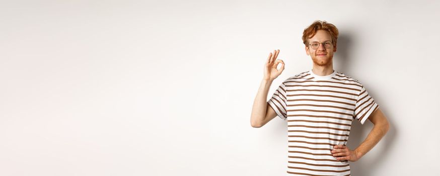 Proud and happy man with red hair and glasses smiling, showing OK sign, praising something excellent, saying yes or good, standing over white background.